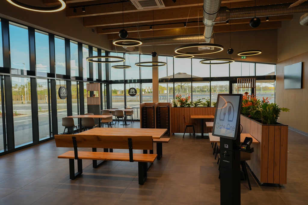 Moderner Café-Bereich mit natürlichem Tageslicht Moderner Café-Bereich mit großen Fenstern, Tischen, Stühlen und Pflanzen, beleuchtet durch kreisförmige Pendelleuchten.