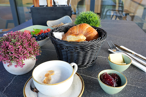 Ein angerichtetes Frühstück auf einem dunklen Esstisch. Zu sehen sind ein Cappuccino mit viel Milchschaum, zwei Croissants in einem Körbchen, Marmelade, Butter und Besteck