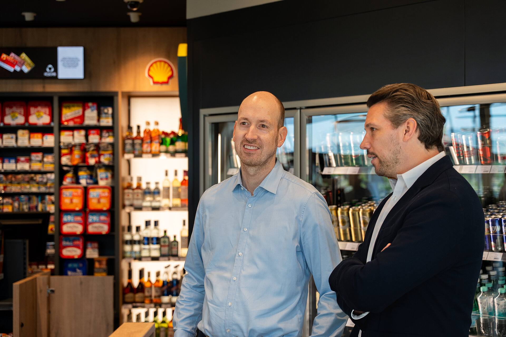 Zwei Personen stehen im Verkaufsbereich eines Tankstellenshops und unterhalten sich miteinander.