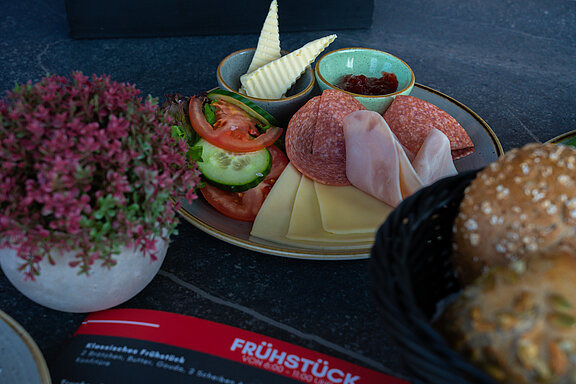 Detailaufnahme von einem Teller Aufschnitt, auf dem Schinken, Salami und Käsescheiben zu sehen sind. Seitlich steht ein geflochtener Brötchenkorb mit verschiedenen Brötchenvariationen.