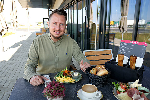 Ein lächelnder Mann sitzt vor einem großzügig gedeckten Frühstückstisch und hält Messer und Gabel in der Hand. Vor ihm steht ein Teller mit frischem Rührei, Tomate und Gurke. Daneben ein geflochtener Korb mit verschiedenen Brötchenvariationen. Vor ihm steht eine Tasse mit frischem Kaffee und daneben ein reichlich gedeckter Teller mit verschiedenen Aufschnitt, Schinken, Salami und Käse
