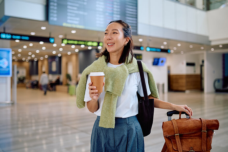 Eine Frau steht mit Koffer und Kaffee in der Hand in einer Flughafenhalle