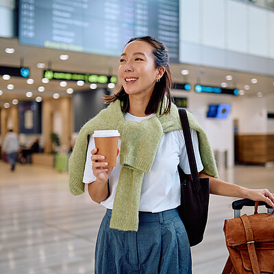 Eine lächelnde Frau steht in einer Flughafenhalle, in einer Hand einen Kaffee to go und in der anderen ihren Koffer