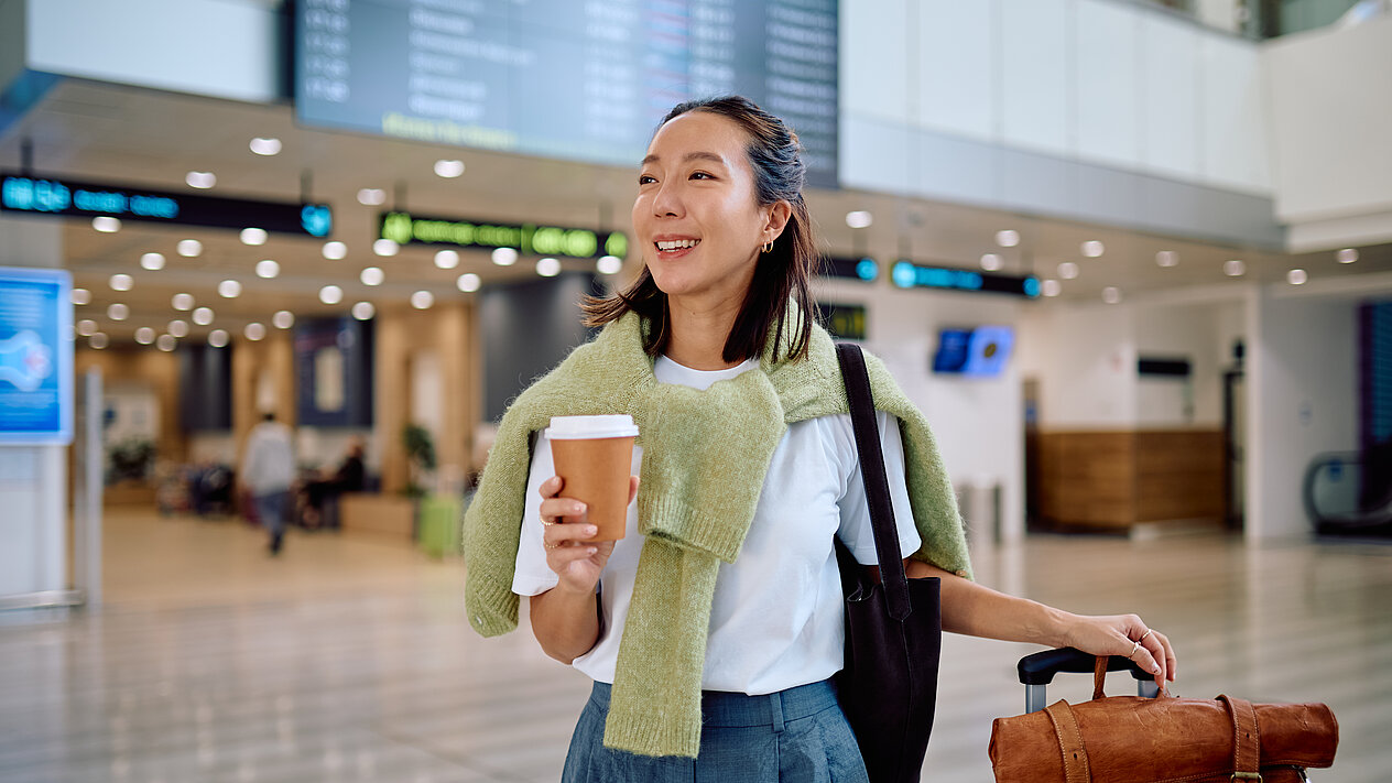 Eine Frau steht am Flughafen mit einem Kaffee in der einen und einem Koffer in der anderen Hand. Hinter ihr ist eine Anzeigetafel von Abflugszeiten der Flugzeugen
