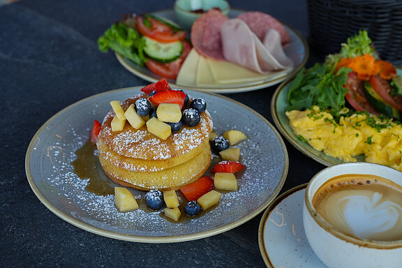 Gedeckter Tisch mit Pancakes, Cappuccino, Rührei und Aufschnitt