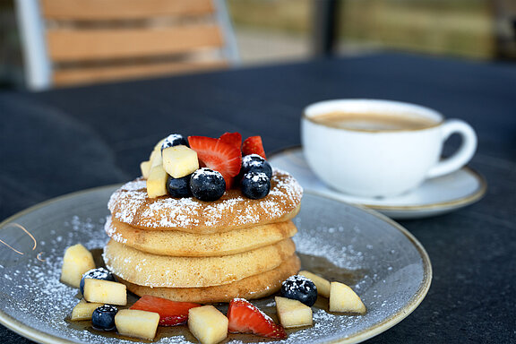 Detailansicht von mehreren aufeinanderliegenden Pancakes, ringsgerum und obendrauf befindet sich frisches Obst und Ahornsirup, dahinter befindet sich eine Tasse frischer, schwarzer Kaffee.