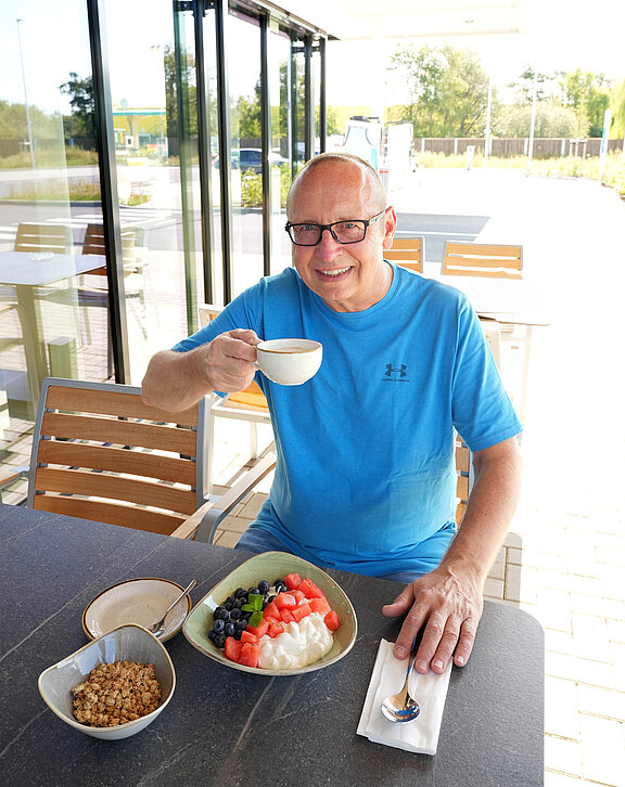 Ein lächelnder Mann sitzt an einem Esstisch mit einer Kaffeetasse in der Hand. Vor ihm steht eine Schüssel Joghurt mit Obst und eine kleine Schüssel Müsli