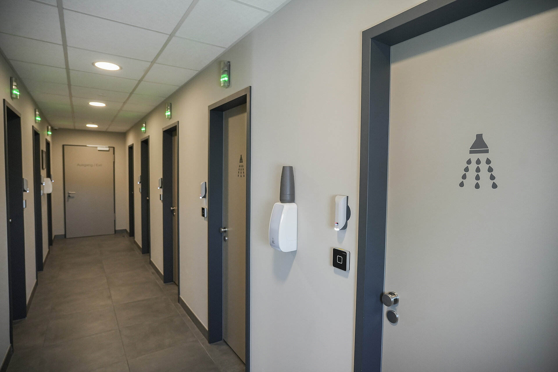 Ein Gang mit vielen Türen auf der linken und rechten Seite, die zu den Duschräumen führen. Über den Türen ist eine Lichtanzeige, die anzeigt, ob die Dusche besetzt oder frei ist.