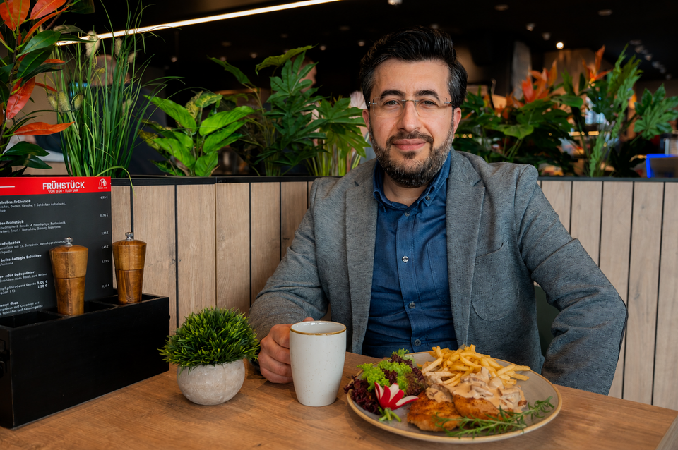 Ein freundlicher Mann schaut in die Kamera und lächelt. Vor ihm ein Schnitzel auf dem Teller, in seiner Hand eine Tasse und er sitzt vor einem Tisch. Auf dem Tisch steht das Menü, auf dem Frühstück zu lesen ist.