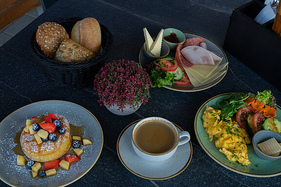 Ein großzügig gedeckter Frühstückstisch mit frischen Pancakes, die mit Ahornsirup und frischem Obst graniert sind. Daneben steht eine Tasse frischer Kaffee und ein geflochtenes Körbchen mit verschiedenen Sorten Brötchen. Außerdem sieht man noch einen gedeckten Teller mit frischem Rührei, Tomaten, Gurken und Salat sowie ein Teller Aufschnitt, auf dem Schinken, Salami und ein paar Scheiben Käse und ein Schälchen Butter zu sehen sind