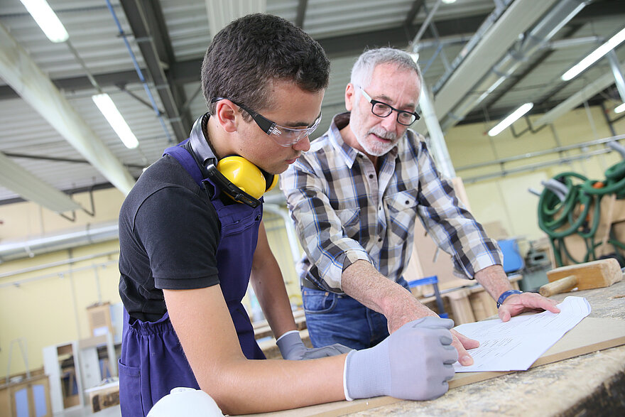 Ein Handwerker und sein Chef stehen an einem Tisch und besprechen Bauzeichungen.