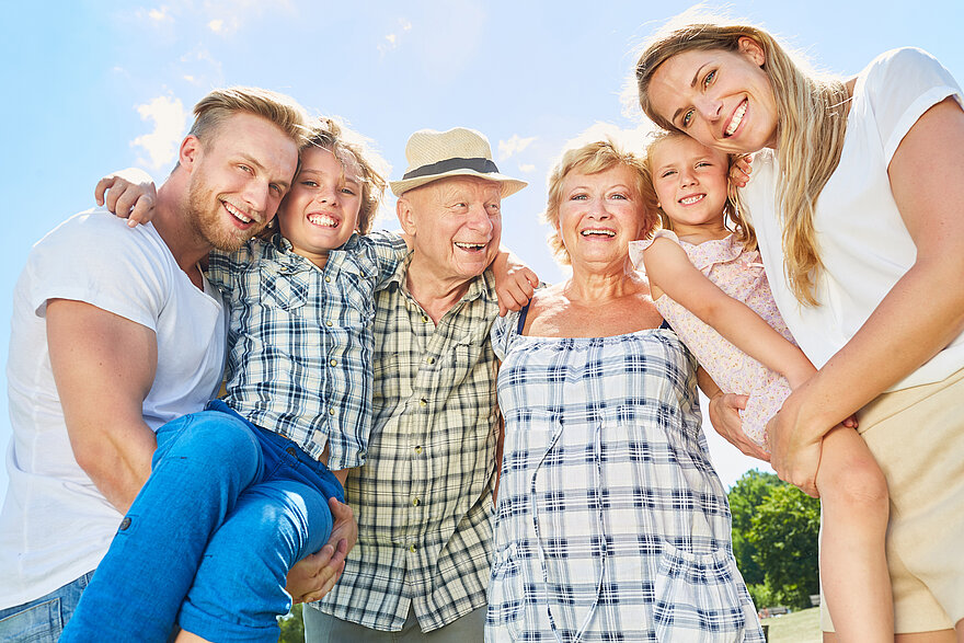 Eine Familie, bestehend aus drei Generationen, steht zusammen und lacht. Darunter Vater, Sohn, Großvater, Großmutter, Tochter und Mutter