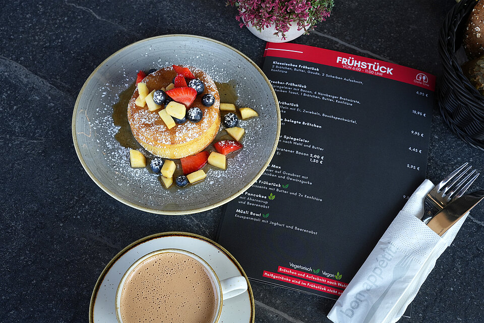 Ein Teller mit frischen, gestalpelten Pancakes, graniert mit frischem Obst, Ahornsirup und Puderzucker, dazu eine Tasse frischer Kaffee und eine Seite der Frühstückskarte mit Preisen