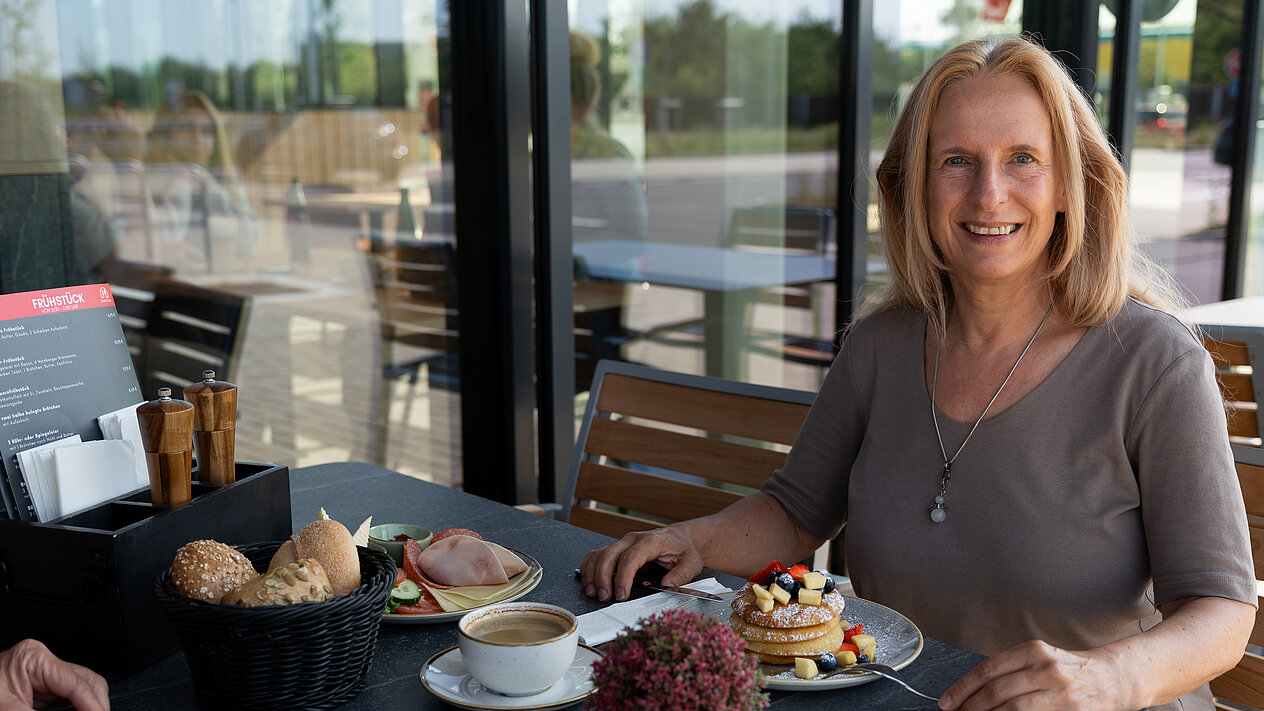 Eine Frau sitzt vor einem reichlich gedeckten Frühstückstisch, vor ihr stehen Rührei, Pancakes mit frischem Obst, ein Brötchenkorb und einem Teller mit Butter und verschiedenem Aufschnitt und Käse