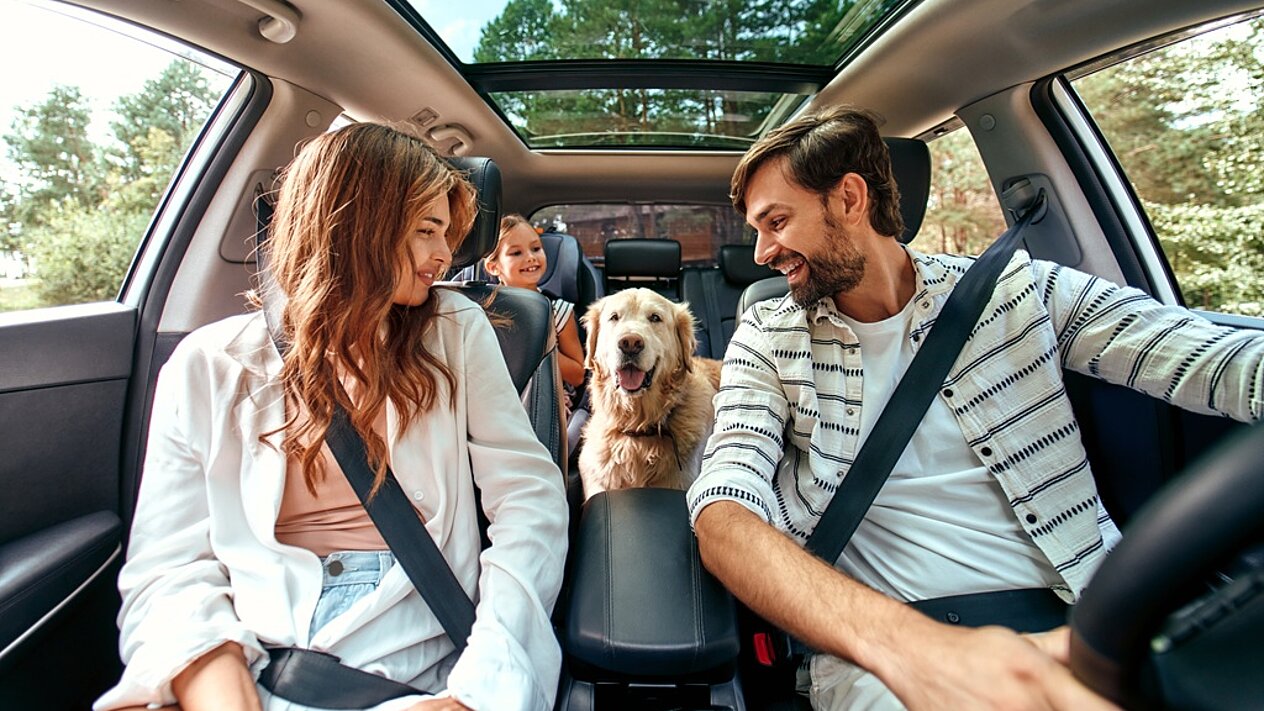 Eine Familie – Vater, Mutter und Kind – sitzt lachend zusammen mit ihrem Golden Retriever in einem modernen Auto mit Panoramadach
