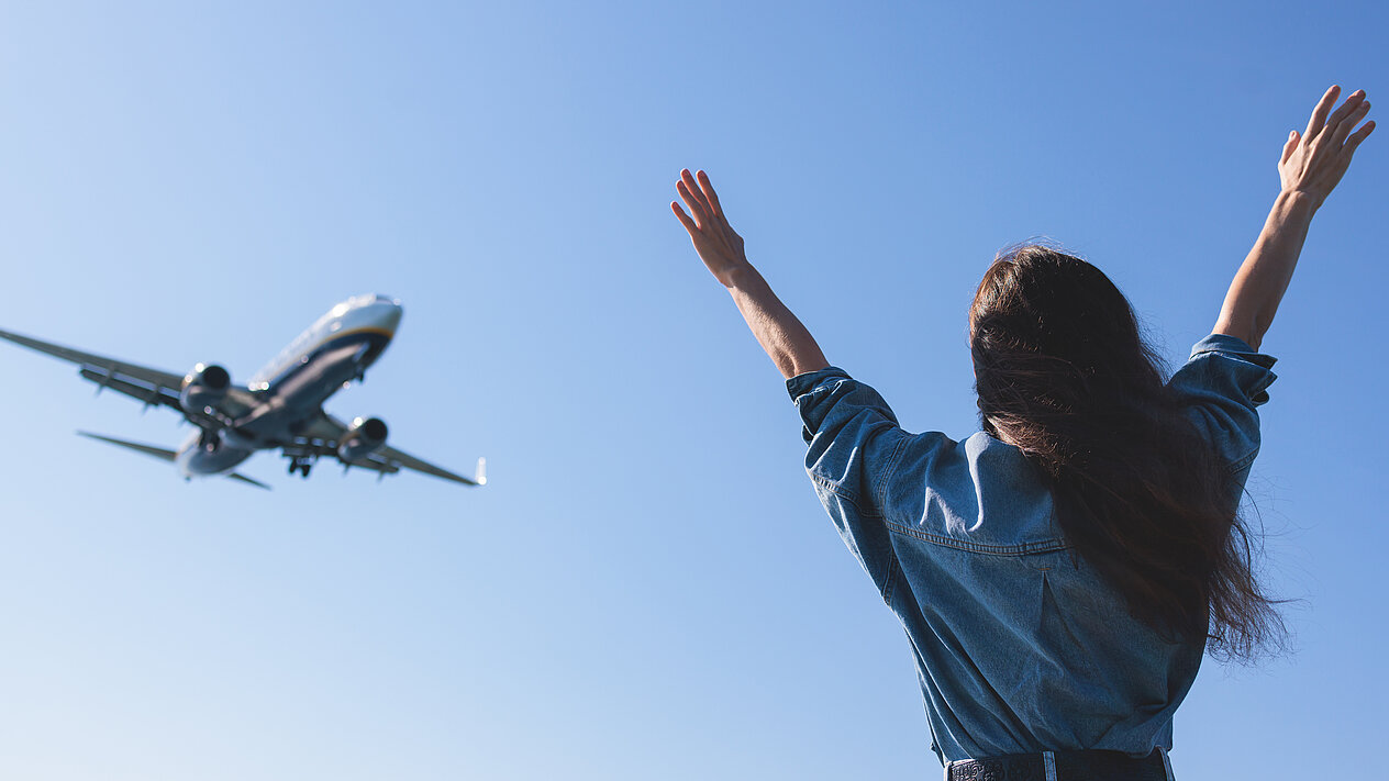 Man sieht eine Frau von hinten, die Hände hochaltend und sich über ein Flugzeug freuend, dass über sie rüberfliegt.