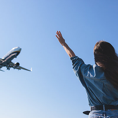 Man sieht eine Frau von hinten, die Arme in die Luft gestreckt und freut sich über ein auf sie zufliegendes Flugzeug. 