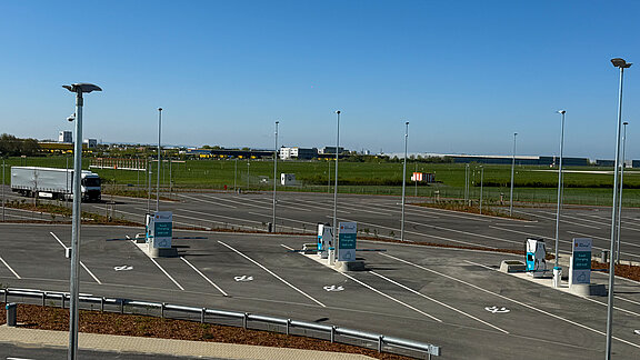 Ein weitläufiger Parkplatz mit mehreren Ladesäulen für Lkw, umgeben von grünen Wiesen und einem klaren blauen Himmel.