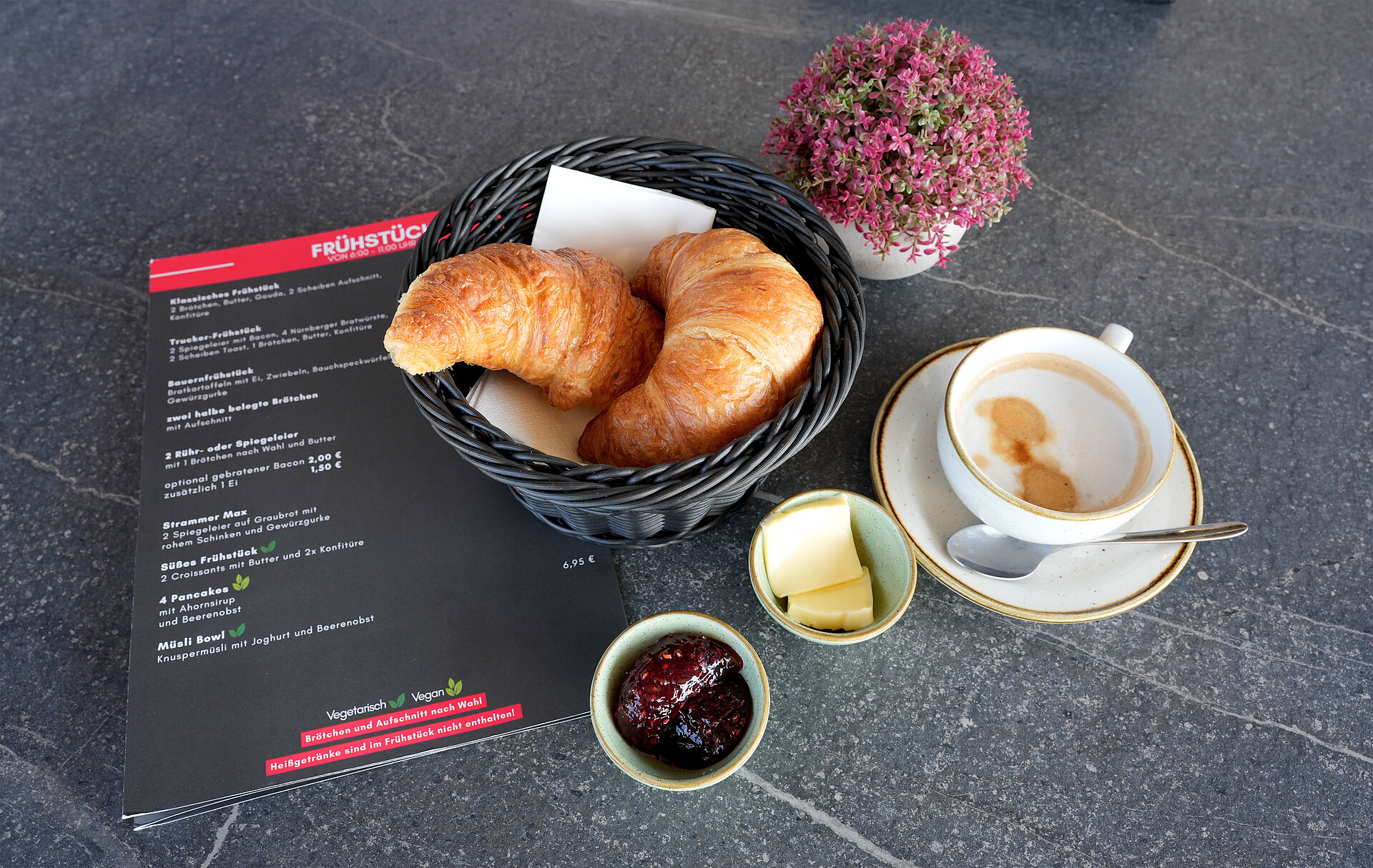Zwei saftige Croissants in einem Frühstückskorb, eine Schale Himbeermarmelade und Butter und eine Tasse Cappuccino auf einem dunklen Esstisch