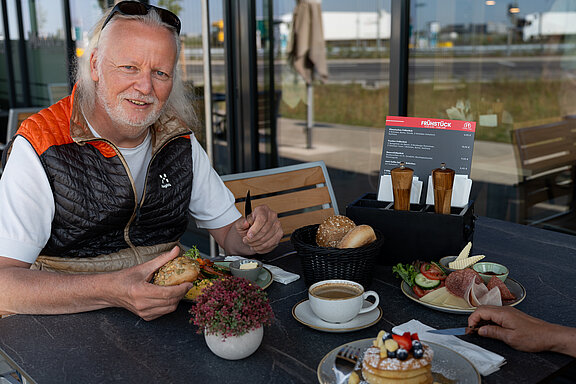 Ein lächelnder Mann sitzt vor einem großzügig gedeckten Frühstückstisch und hält ein Brötchen und ein Messer in der Hand. Vor ihm steht eine frische Tasse Kaffe und gegenüber frische Pancakes mit Ahornsirup und Obst.