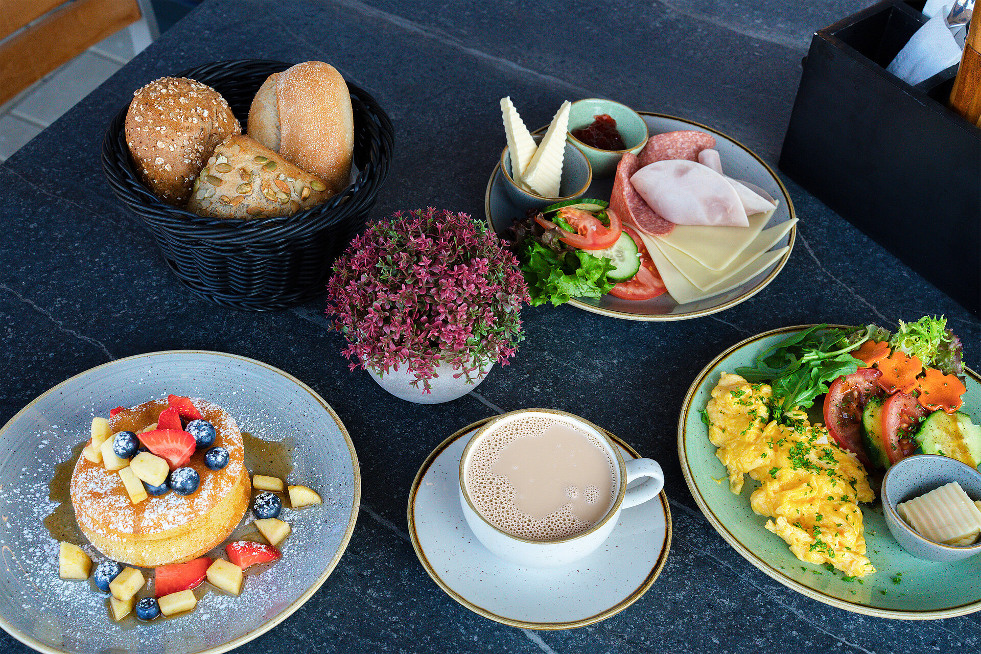 Gedeckter Frühstückstisch mit Pancakes, Kaffee, Rührei, Brötchenvariationen, ein Teller mit Aufschnitt und Käse, Tomaten, Gurken und Salat