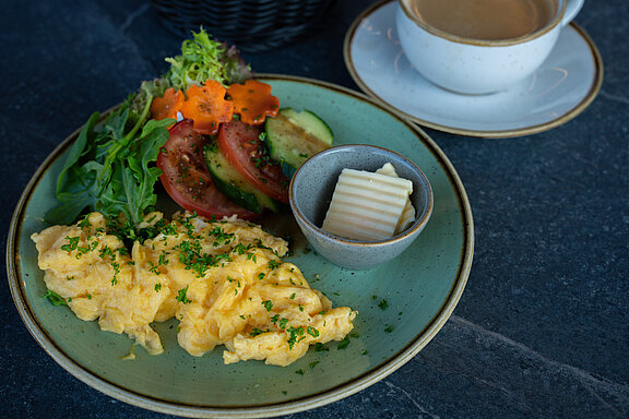 Detailansicht von frisch zubereiteten Rührei mit Schnittlauch, Tomaten, Gurken und Salat auf einem großen Teller mit einem Schälchen Butter. Dahinter befindet sich eine Tasse frischer, heißer Kaffee